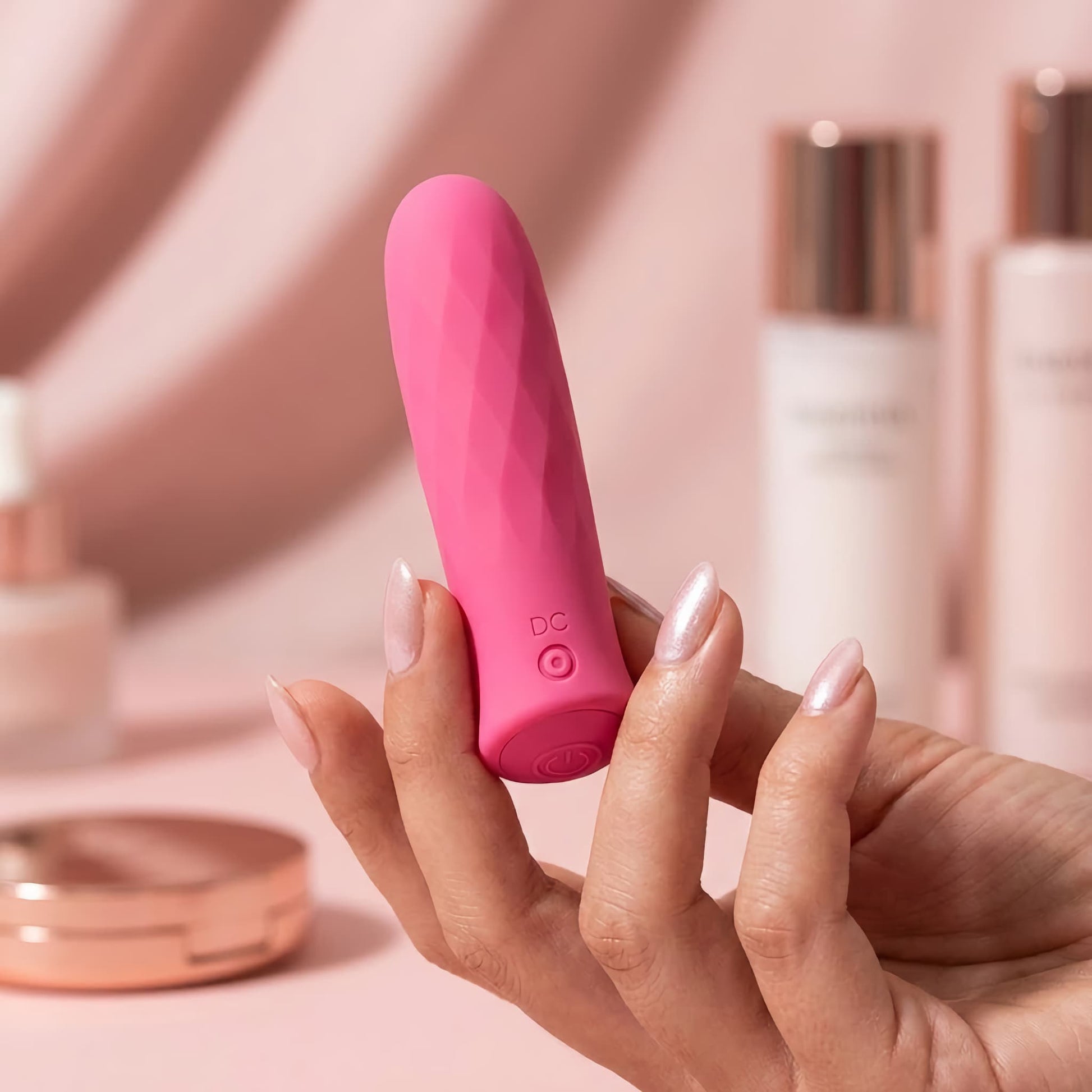 Close-up shot of a woman holding a small pink faceted bullet vibrator between her fingertips, displayed alongside elegant skincare bottles.