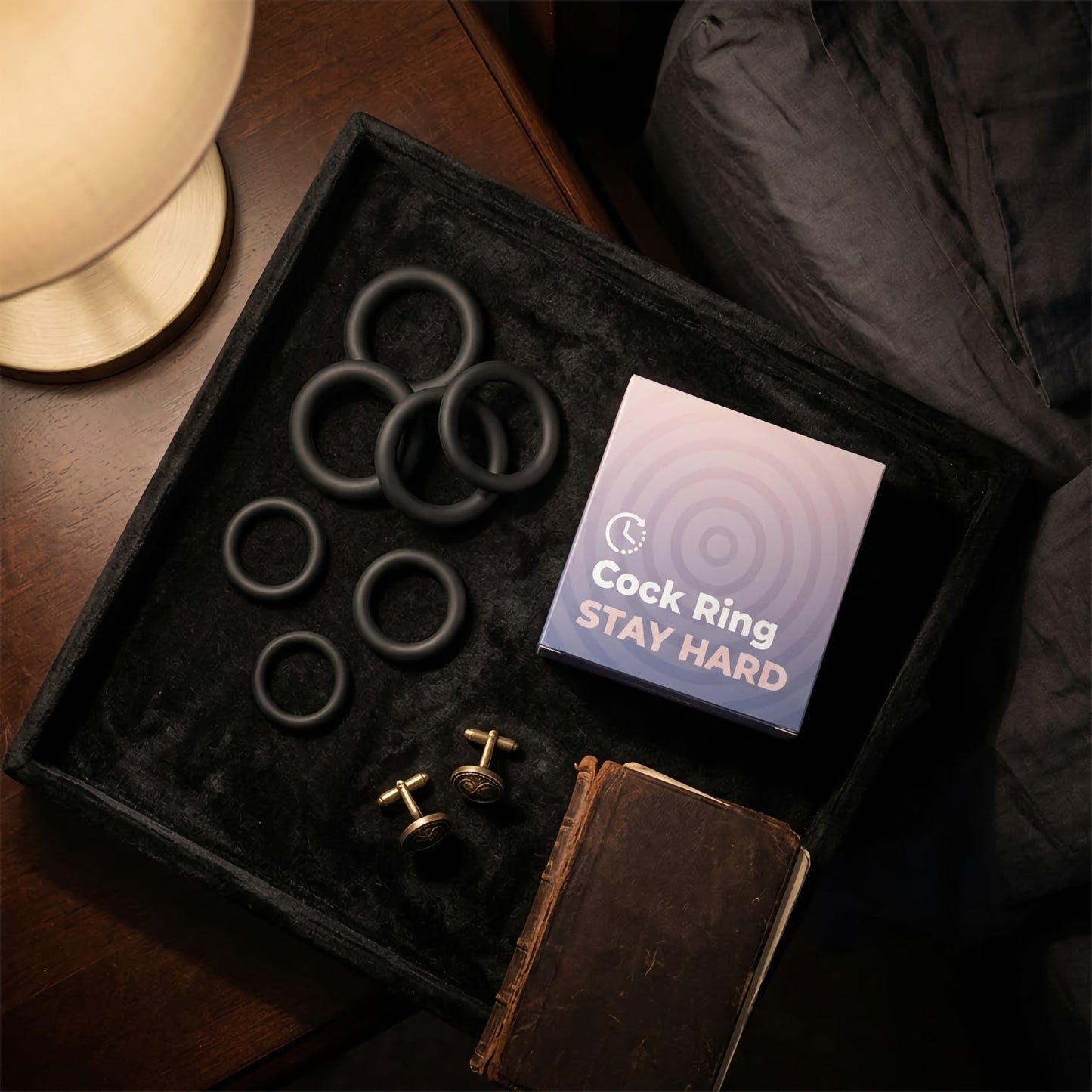 Silicone cock ring set displayed on a velvet valet tray with its 'Stay Hard' product box, antique gold cufflinks, and a leather notebook on a dark wooden nightstand.