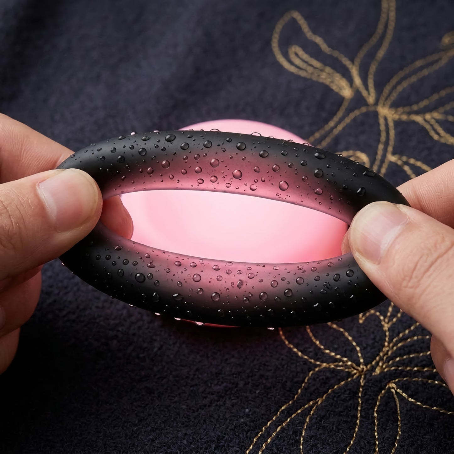 Flexible and waterproof black silicone cock ring being gently stretched. Covered in delicate water droplets on a dark textured fabric with gold embroidery.
